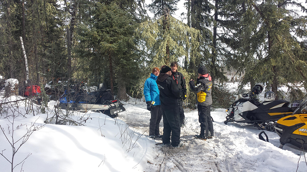 A group of people are standing and taking a break from snowmobiling in the boreal forest in northern Saskatchewan