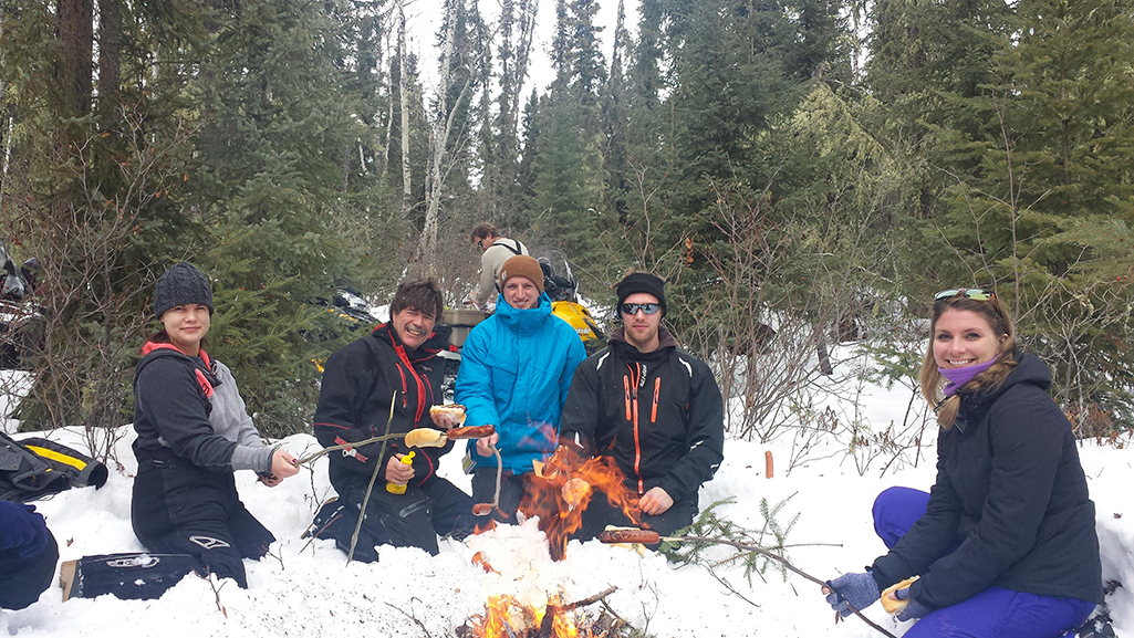 A group of people are gathered around a snowy wintertime fire in the forest while taking a break from snowmobiling in northern Saskatchewan