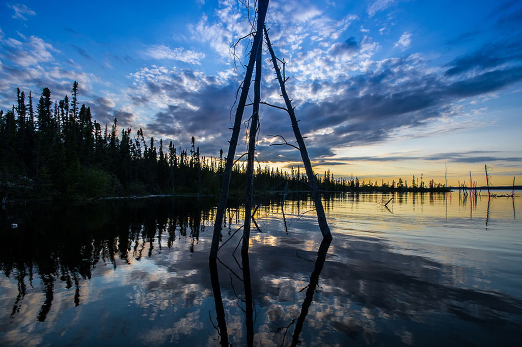 The sun sets on a beautiful evening in Tazin Lake, Saskatchewan.  A structure constructed with three logs resembles a tipi sits in the water.